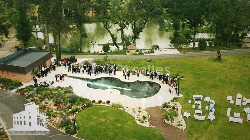 Vue aérienne d'un jardin avec piscine entourée d'invités, près d'un bâtiment et d'un parc arboré avec étang.