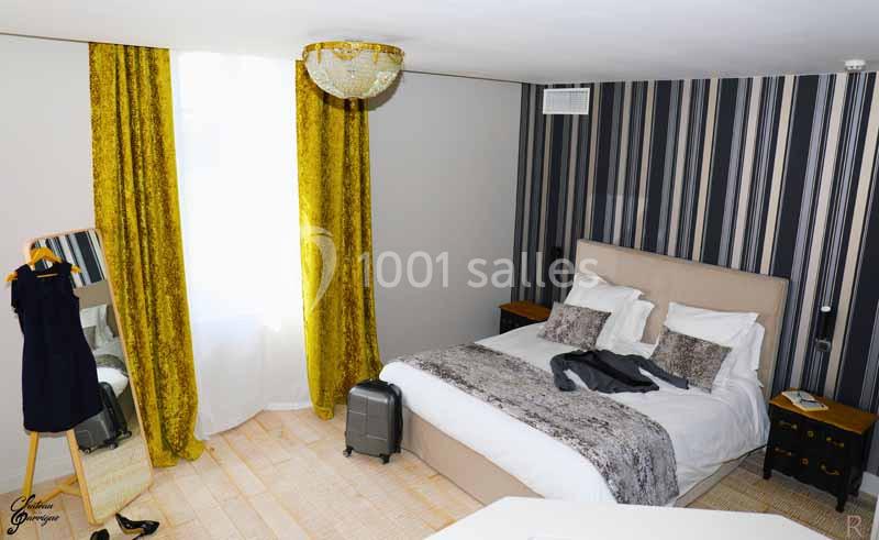 Chambre lumineuse avec lit double, rideaux jaunes, valise au sol et robe noire sur un miroir sur pied.