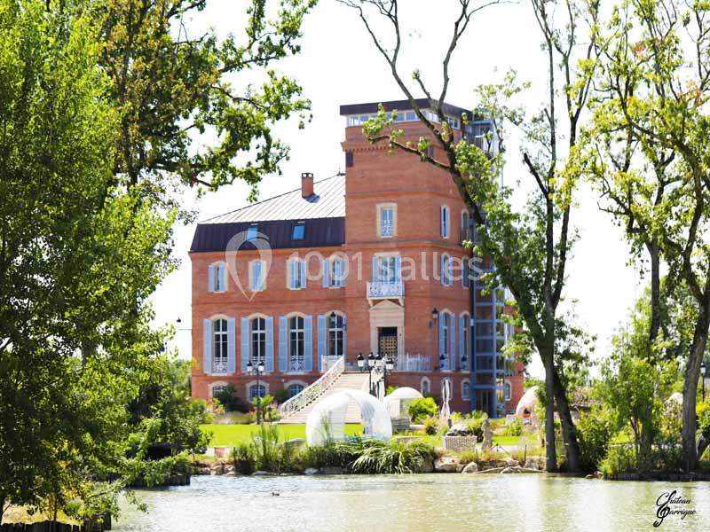 Château en briques rouges entouré d'arbres, avec un étang au premier plan et un ciel dégagé en arrière-plan.