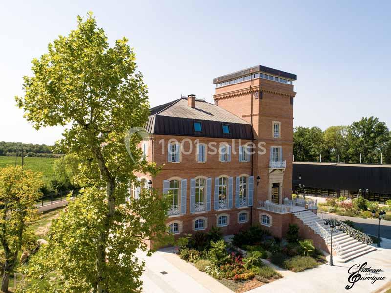 Vue aérienne d'un château en briques rouges entouré de jardins, avec des champs et des arbres en arrière-plan.