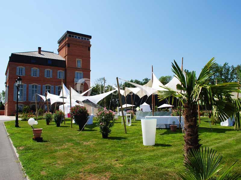 Jardin avec pelouse, palmiers, tentes blanches et un bâtiment en briques rouges sous un ciel dégagé.