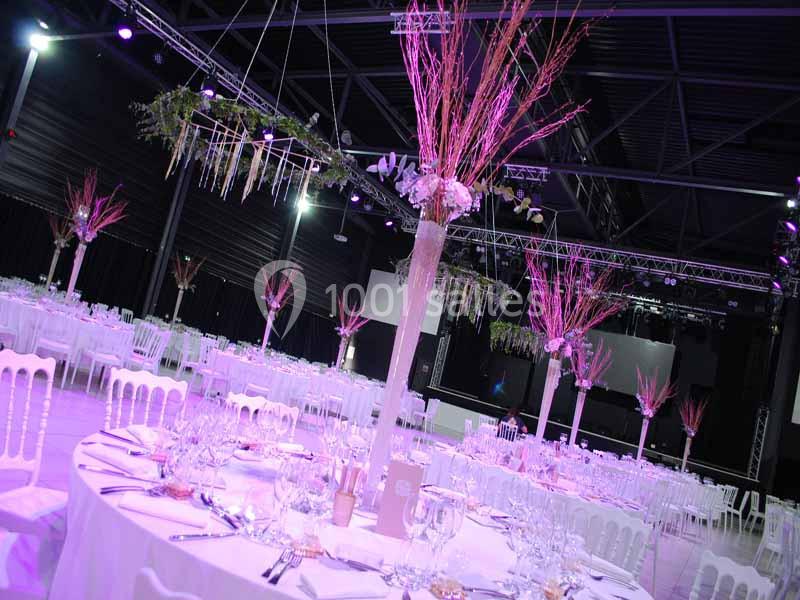 Salle de réception décorée avec des tables rondes, nappes blanches, et centres de table fleuris illuminés en rose.