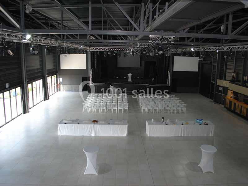 Salle spacieuse avec scène, rangées de chaises blanches, tables dressées et éclairage industriel moderne.