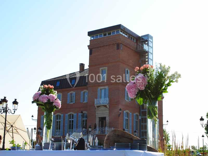 Grande maison en briques rouges avec une tour, vue depuis une table dressée avec des bouquets de fleurs au premier plan.