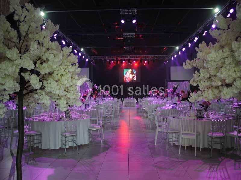 Salle de réception décorée avec des tables rondes, des chaises blanches et des arbres fleuris sous un éclairage coloré.