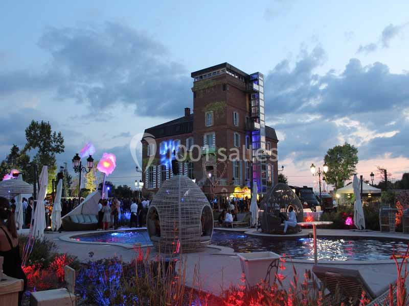 Fête en soirée près d'un bâtiment éclairé, avec des installations lumineuses et un bassin entouré de végétation.