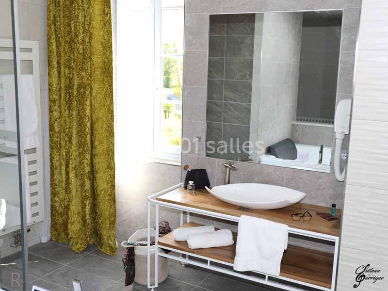 Salle de bain lumineuse avec lavabo sur meuble en bois, miroir mural, rideau jaune et fenêtre donnant sur l'extérieur.