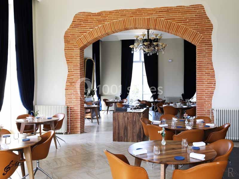 Salle de restaurant lumineuse avec tables en bois, chaises orange et arche en briques rouges.