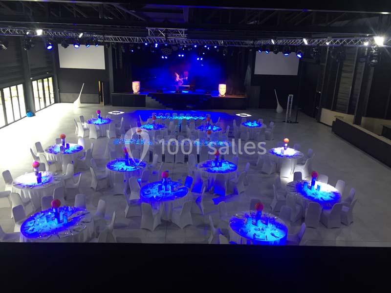 Salle de réception avec tables rondes décorées et éclairage bleu, scène en fond avec projecteurs allumés.