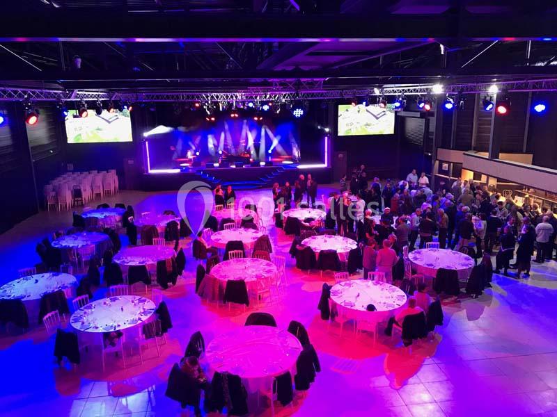 Salle de réception avec tables rondes dressées, éclairage coloré et scène en arrière-plan lors d'un événement.