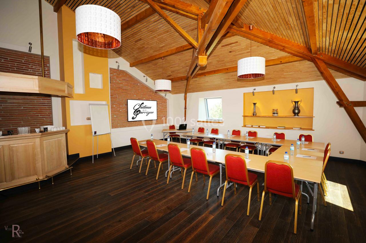 Salle de réunion lumineuse avec poutres apparentes, tables disposées en U, chaises rouges et écran mural.