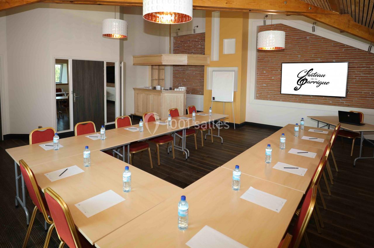 Salle de réunion lumineuse avec tables disposées en U, chaises rouges, bouteilles d'eau et écran de présentation.