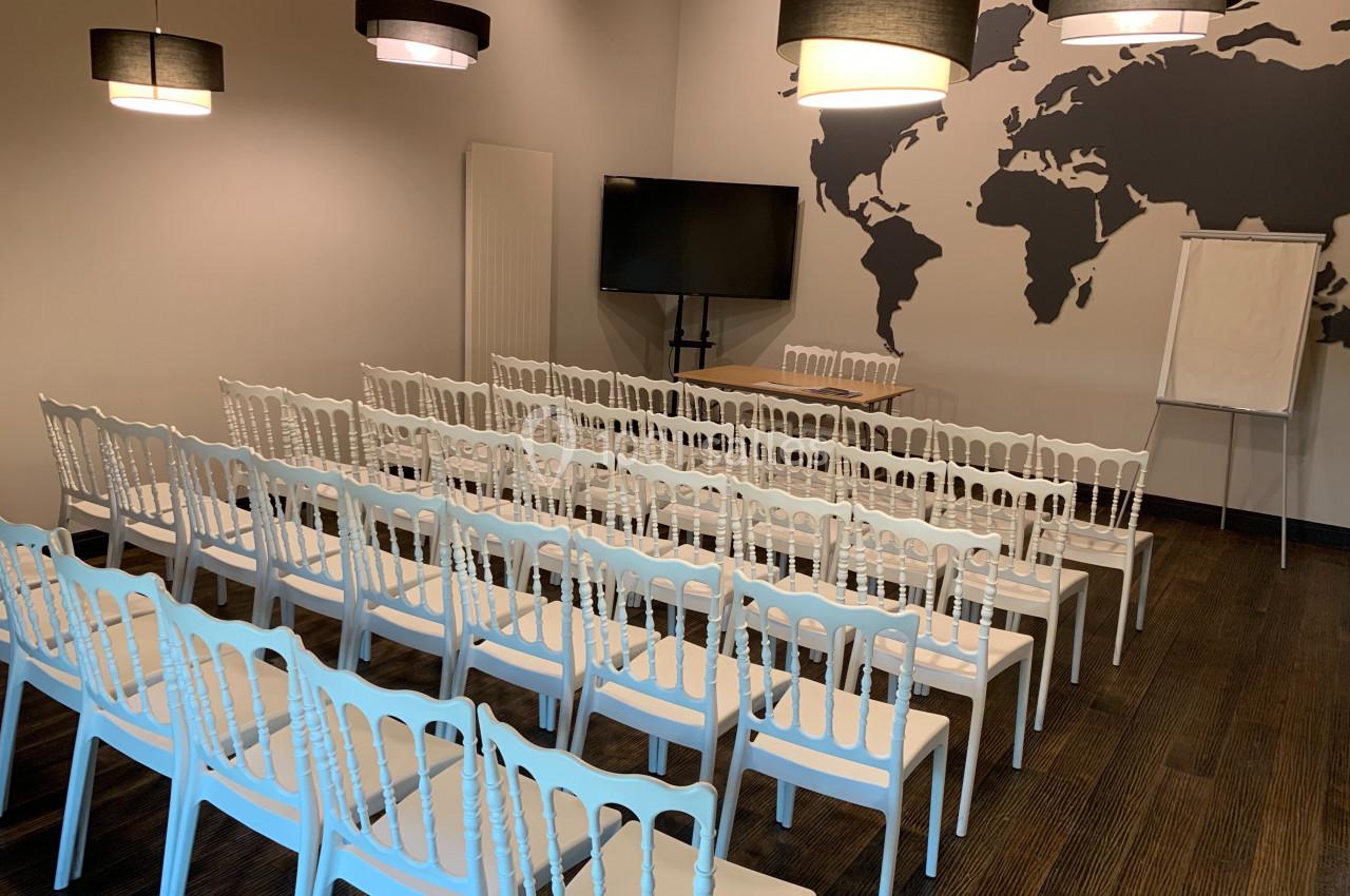 Salle de réunion avec des rangées de chaises blanches, un écran, un tableau blanc et une carte du monde au mur.