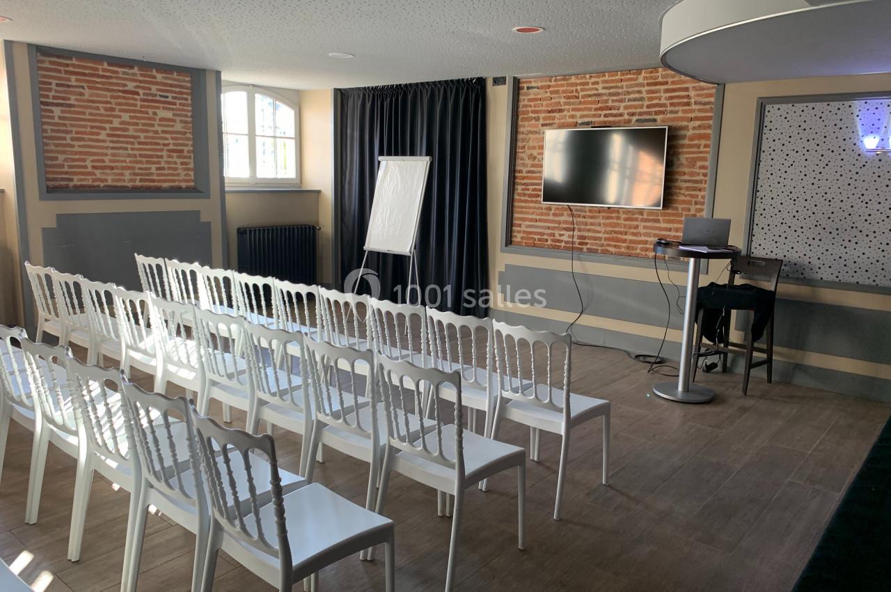 Salle de réunion lumineuse avec chaises blanches alignées, écran, chevalet de présentation et murs en briques apparentes.