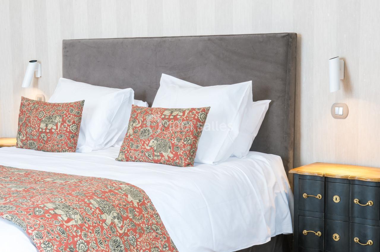 Lit double avec tête de lit grise, draps blancs et coussins décoratifs à motifs rouges, encadré par deux tables de chevet.