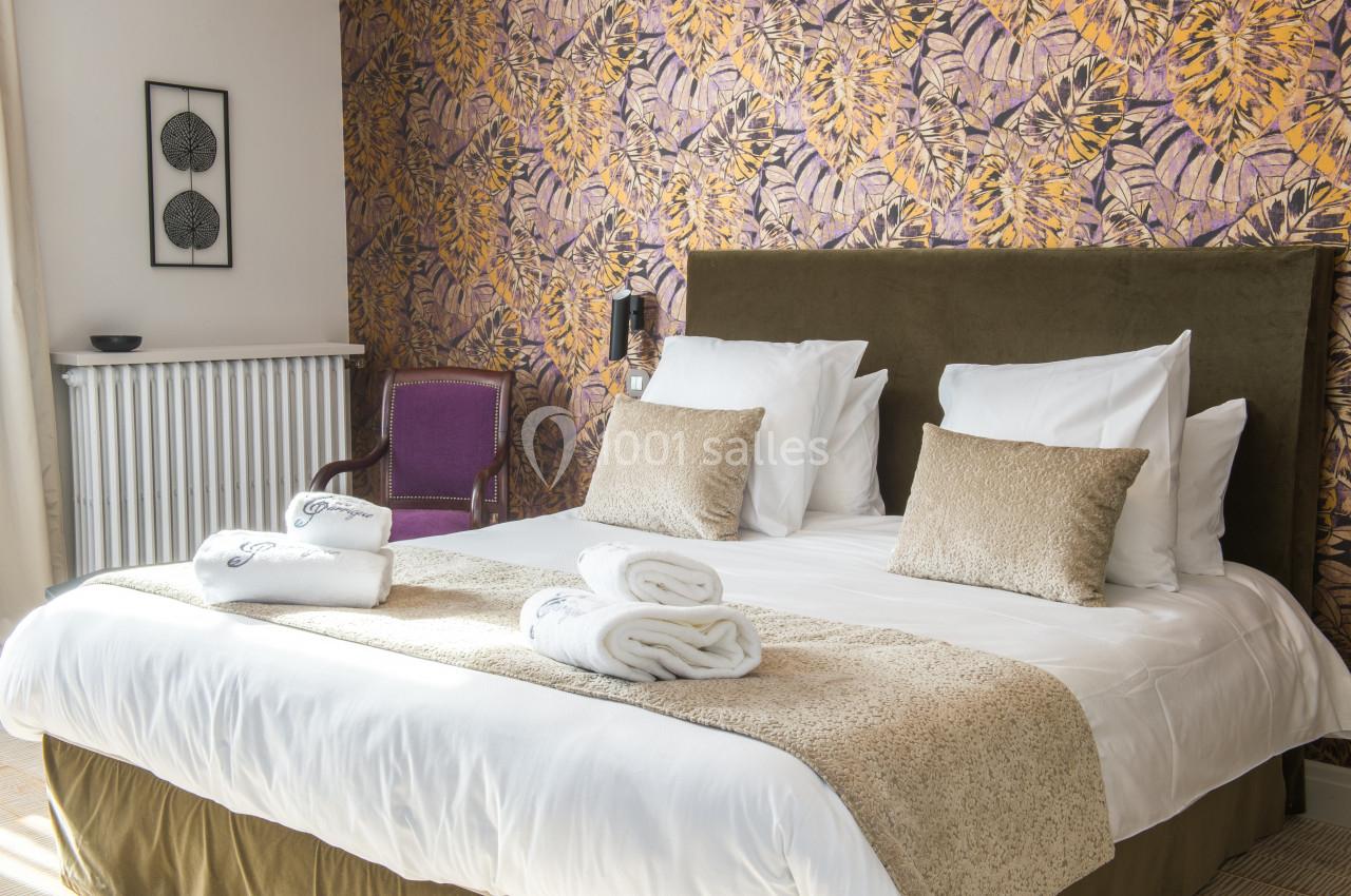 Lit double avec coussins et serviettes pliées, devant un mur à motifs floraux et une chaise violette.