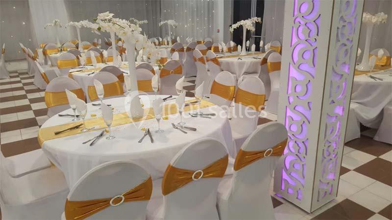Salle de réception décorée avec des tables rondes, nappes blanches, chaises ornées de rubans dorés et centres de table…