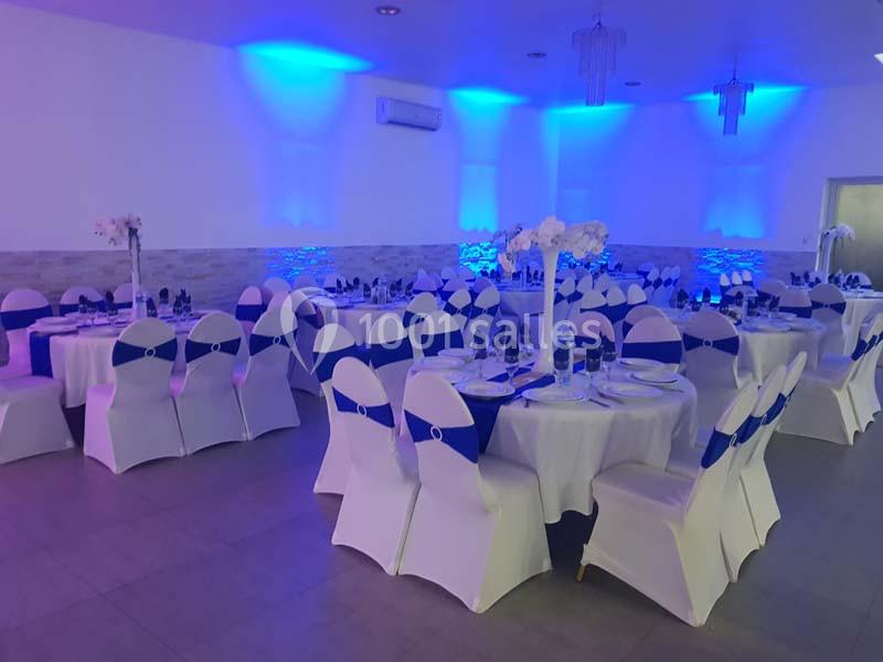 Salle de réception décorée avec des tables rondes, nappes blanches, chaises couvertes et éclairage bleu tamisé.