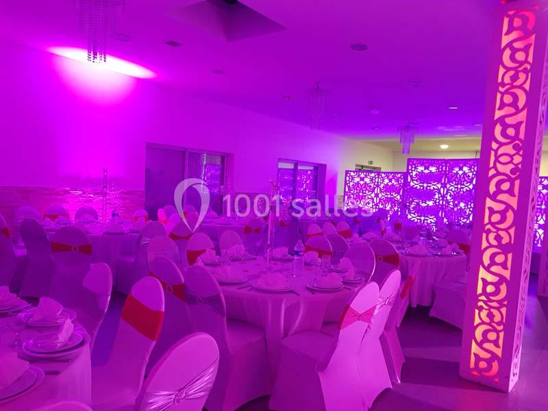 Salle de réception décorée avec des tables rondes dressées, chaises blanches et éclairage rose vif.