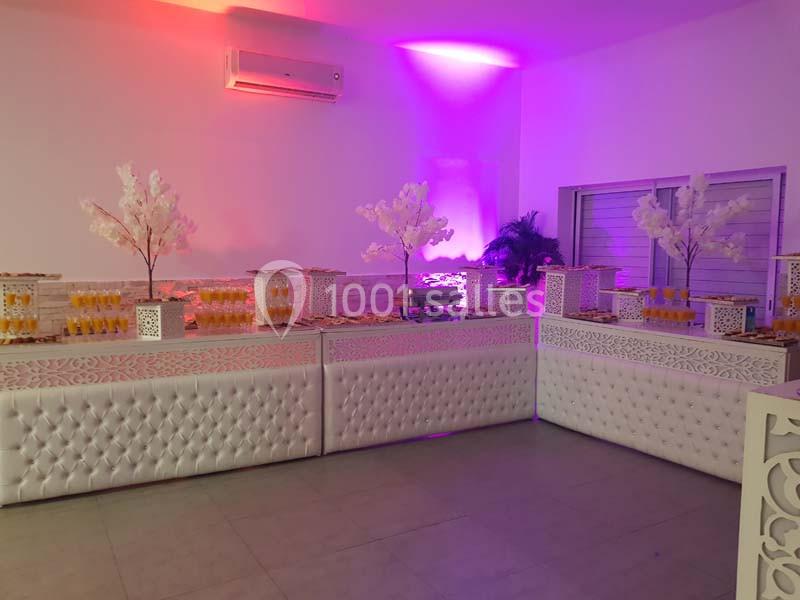 Buffet décoré avec des tables blanches capitonnées, des arbres artificiels et des boissons alignées dans une salle éclairée.