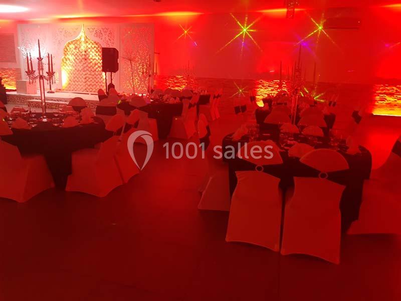 Salle de réception décorée avec des tables dressées, chaises blanches et noires, éclairage rouge et ambiance festive.
