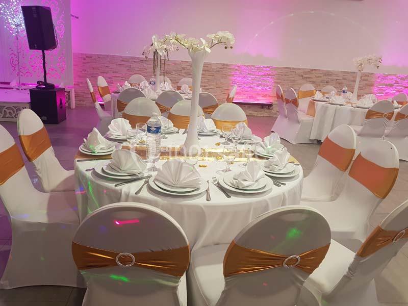 Table ronde décorée pour un événement, avec nappes blanches, serviettes pliées, chaises ornées de rubans dorés et centre de…