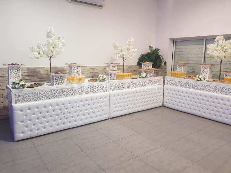 Buffet décoré avec des tables blanches capitonnées, boissons et arbres artificiels ornés de fleurs blanches.