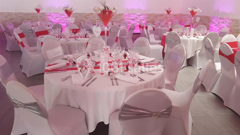Salle de réception décorée avec des tables rondes, nappes blanches, chaises habillées et accents rouges.
