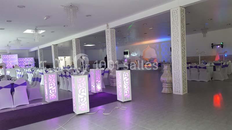 Salle de réception décorée avec des tables dressées, des chaises blanches ornées de nœuds violets et un éclairage coloré.