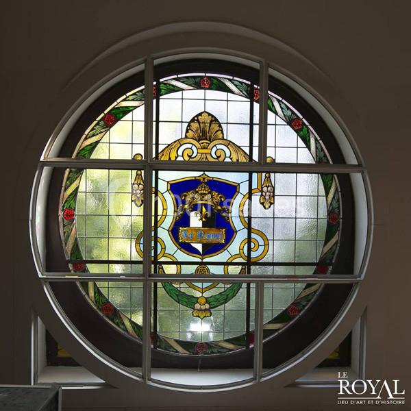 Vitrail rond coloré avec un blason central bleu et doré, encadré par une structure métallique blanche.