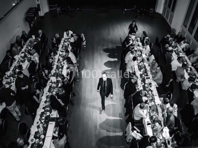 Un homme en costume marche au centre d'une salle où des convives sont assis à de longues tables lors d'un événement.