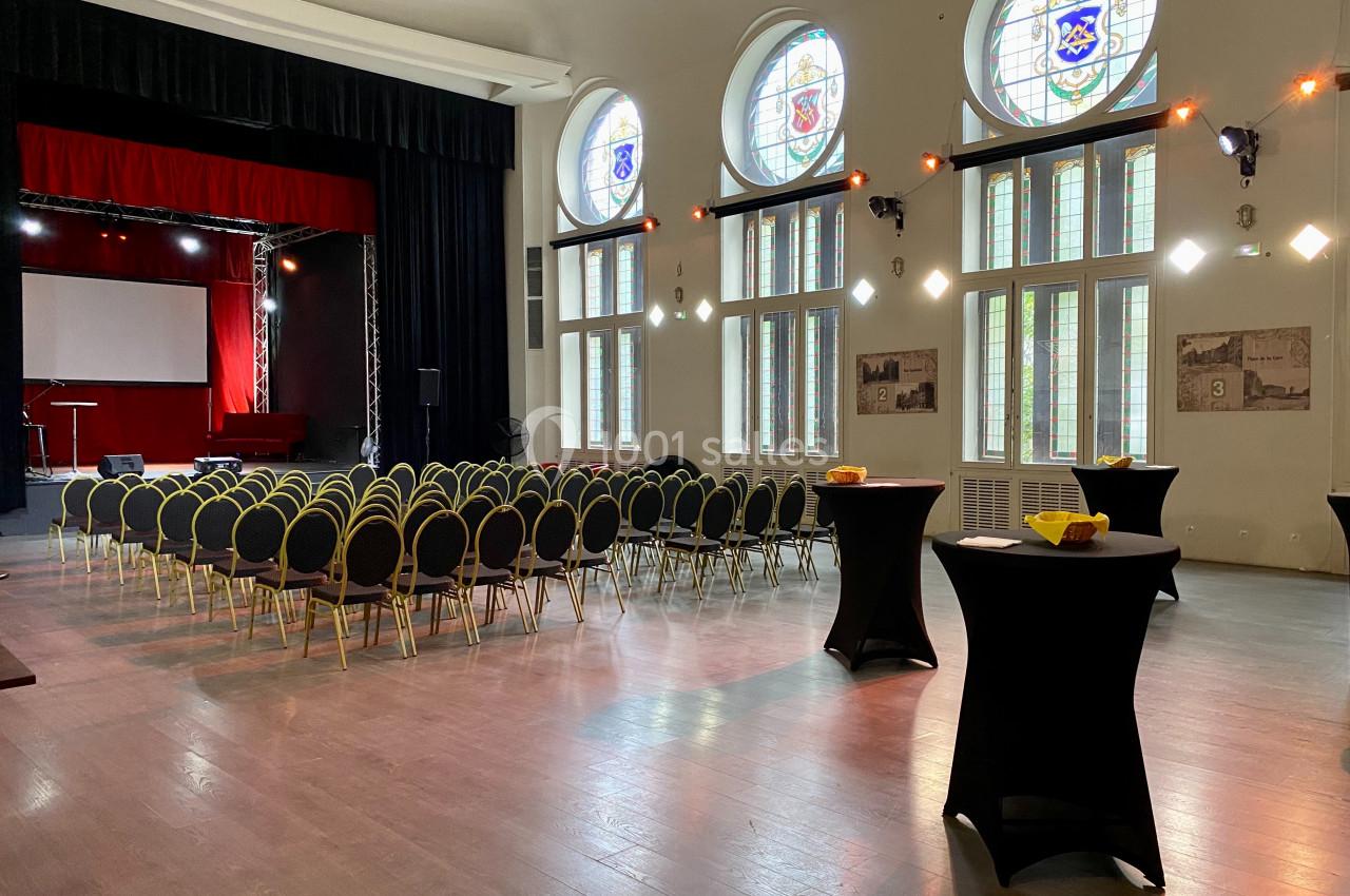 Salle de réception lumineuse avec des rangées de chaises, une scène équipée d'un écran et des tables hautes en premier plan.