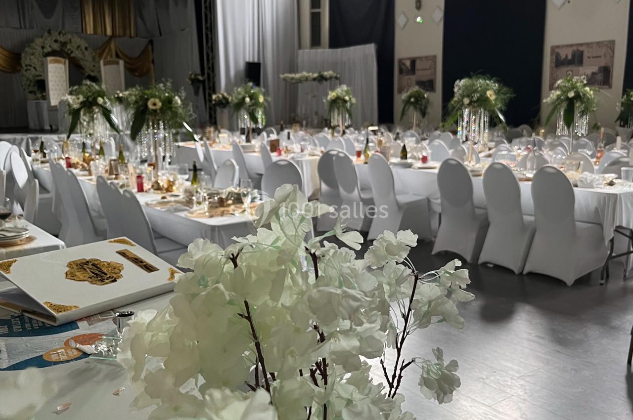 Salle de réception décorée pour un événement, avec tables dressées, chaises blanches et arrangements floraux élégants.