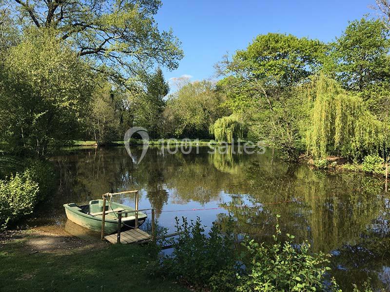 Barque amarrée sur un petit ponton au bord d'un étang entouré d'arbres et de végétation sous un ciel bleu.