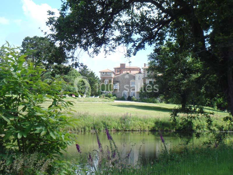 Vue d'un château en pierre entouré de verdure, avec un étang au premier plan et des arbres encadrant la scène.