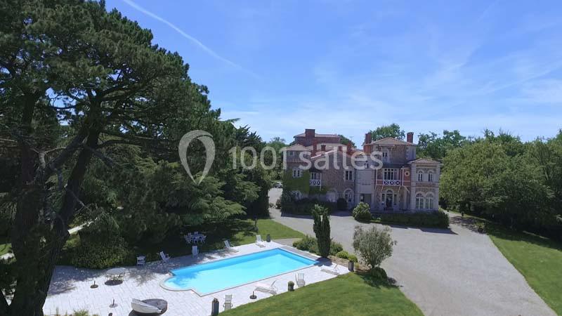Vue aérienne d'une grande maison en pierre avec piscine, entourée de jardins et d'arbres sous un ciel dégagé.