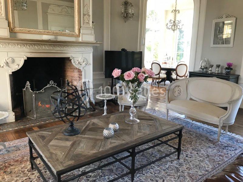 Salon élégant avec cheminée allumée, mobilier classique, table basse en bois et vase de roses sur un tapis décoratif.