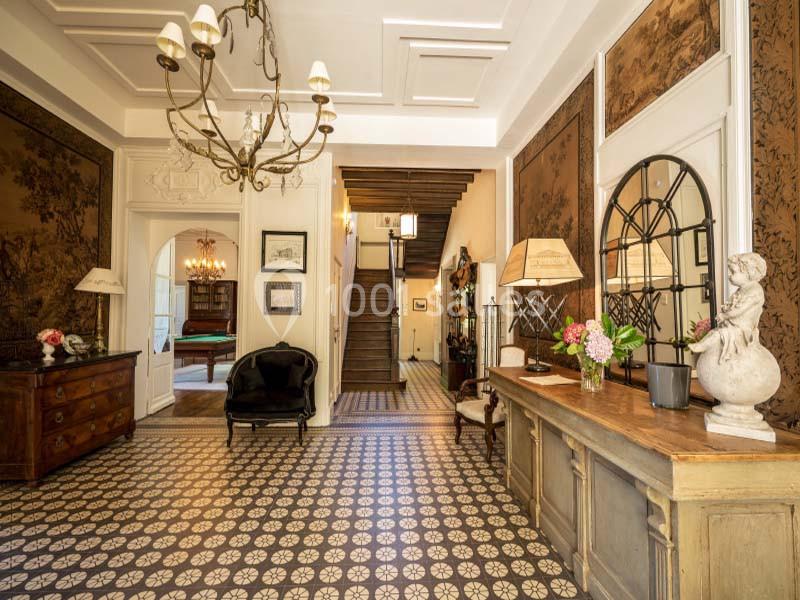 Entrée spacieuse avec carrelage à motifs, meubles anciens, lustre et escalier en bois au fond.