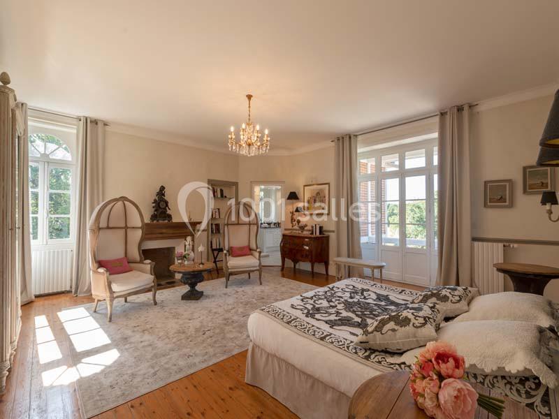 Chambre lumineuse avec lit double, fauteuils, cheminée, meubles anciens et grandes fenêtres donnant sur un jardin.