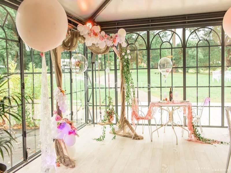 Décoration romantique avec ballons et fleurs dans une verrière, table dressée pour deux avec vue sur un jardin.