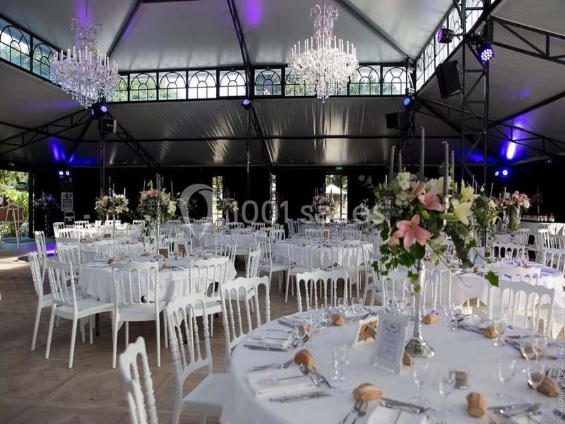 Salle de réception élégante avec tables dressées, chaises blanches, chandeliers suspendus et décorations florales.