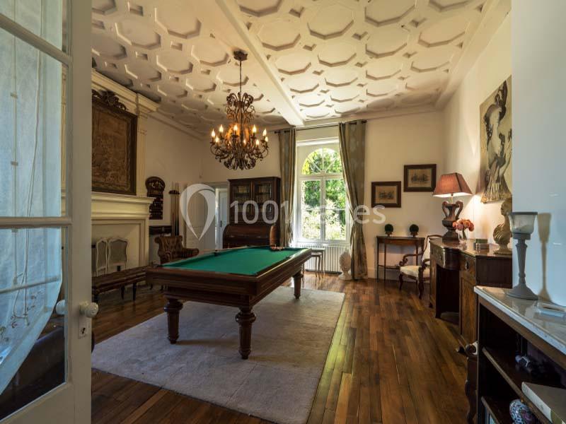 Salle élégante avec table de billard, parquet en bois, lustre, cheminée et grande fenêtre donnant sur un jardin.