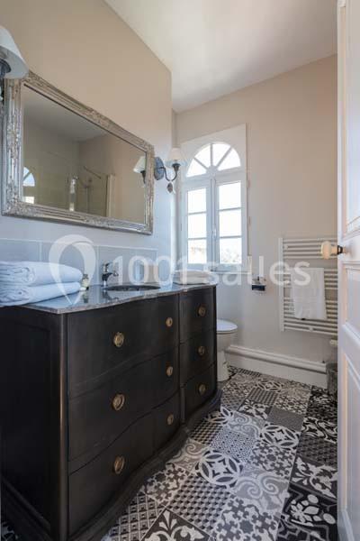 Salle de bain avec meuble en bois sombre, miroir encadré, fenêtre cintrée et sol en carreaux à motifs.