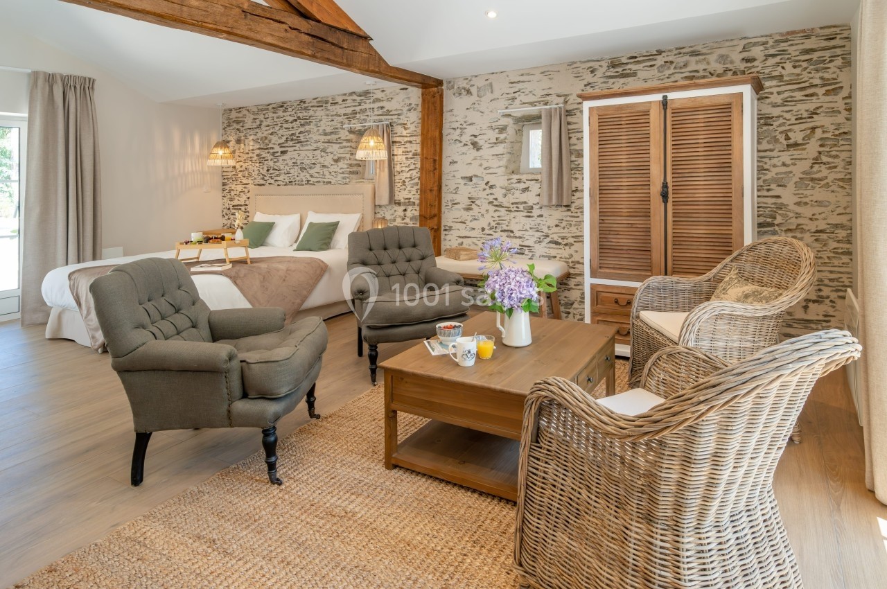 Chambre lumineuse avec murs en pierre, lit double, fauteuils, table basse et armoire en bois dans un décor rustique.