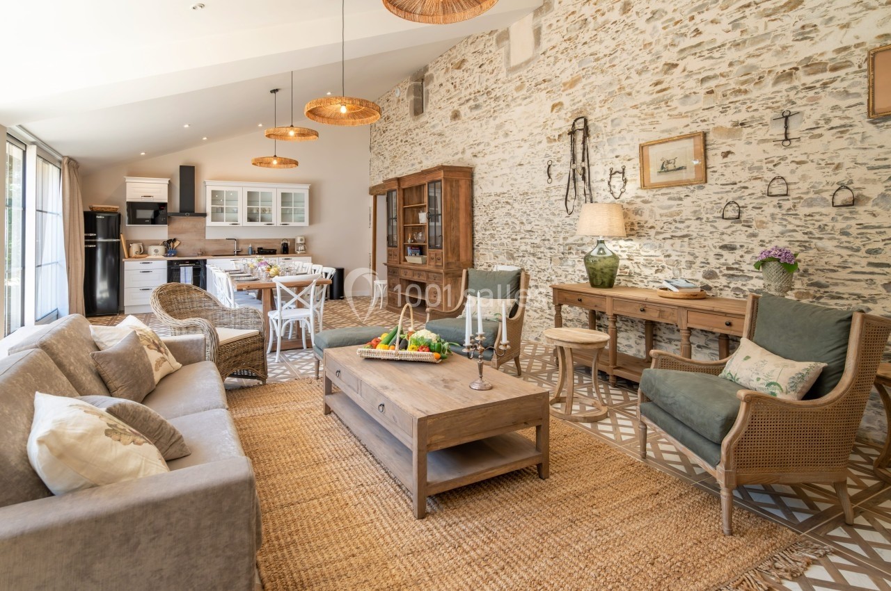 Salon lumineux avec murs en pierre, mobilier en bois, canapé beige, et cuisine ouverte en arrière-plan.