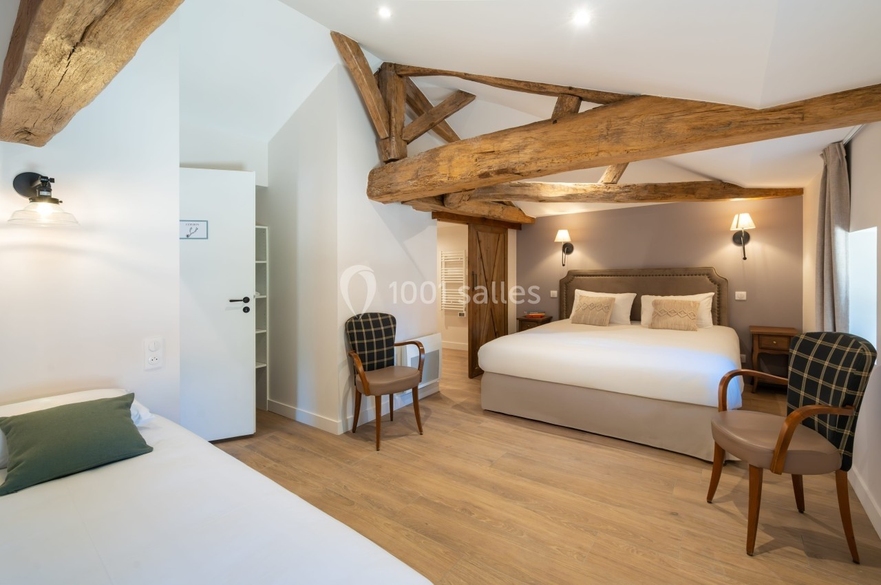 Chambre lumineuse avec poutres apparentes, lit double, lit simple, deux fauteuils et éclairage chaleureux.