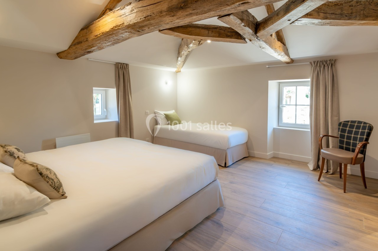 Chambre lumineuse avec poutres apparentes, deux lits, sol en bois clair, deux fenêtres et une chaise près du mur.