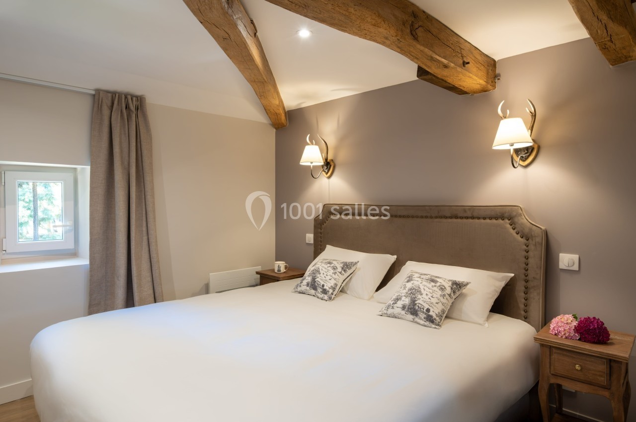 Chambre lumineuse avec lit double, poutres apparentes, murs clairs, lampes murales et table de chevet en bois.