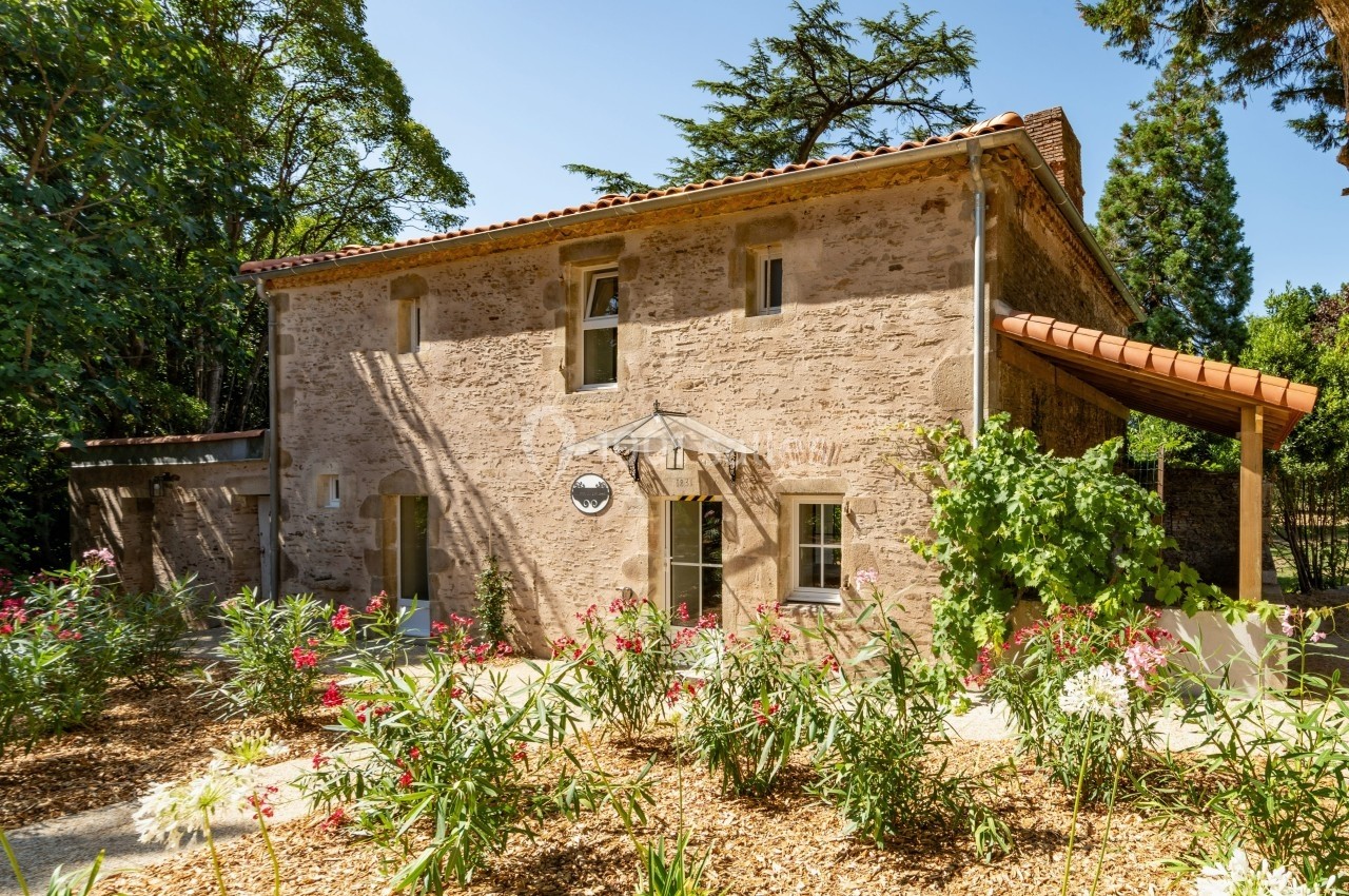 Maison en pierre avec toit en tuiles, entourée de végétation et de fleurs, sous un ciel dégagé.