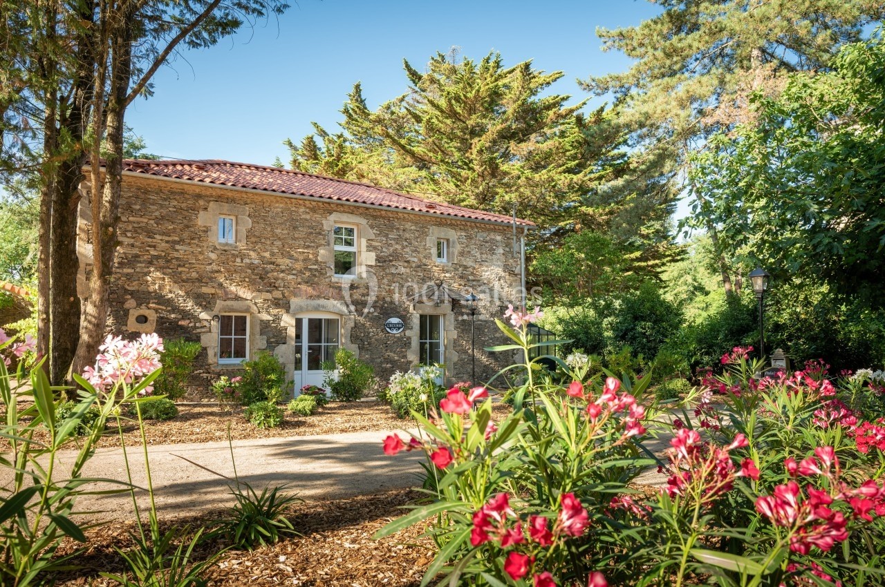 Maison en pierre entourée de végétation luxuriante et de fleurs colorées sous un ciel bleu clair.